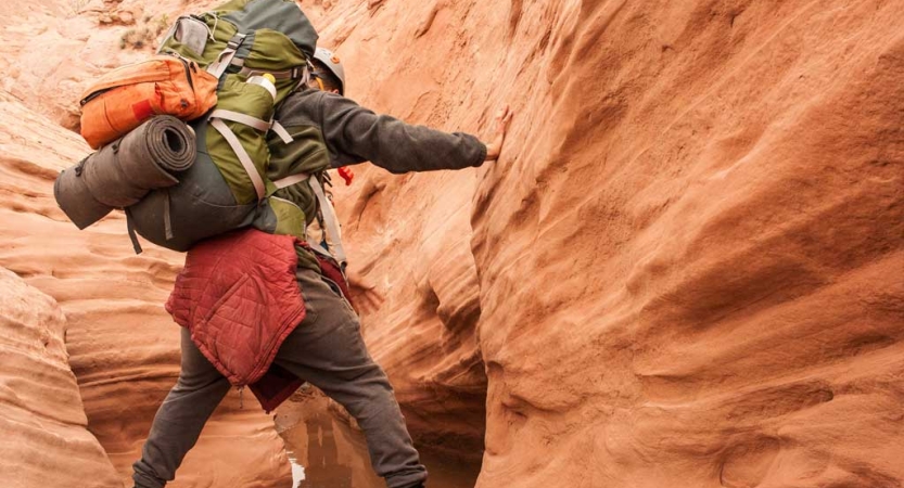 A person wearing a backpack maneuvers through a red slot canyon 